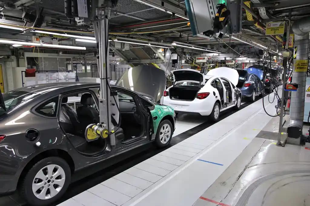 Car bodies on a factory assembly line, a dark hatchback with its doors and bonnet open in the foreground and a row of white and blue bodies receding down the line behind it, under overhead conveyor gantries.