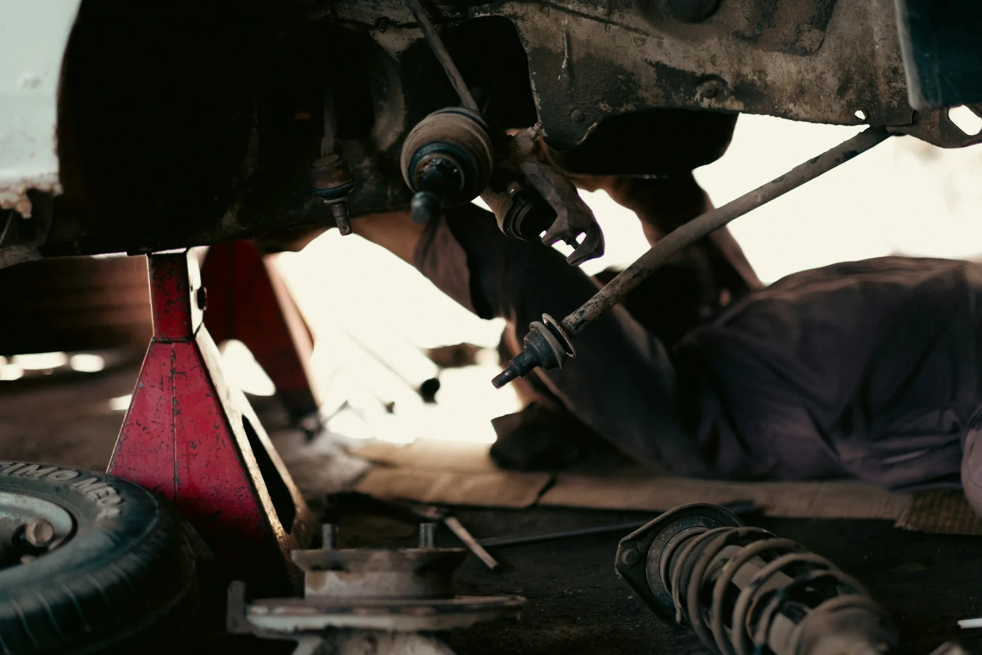 A car raised on a red axle stand in a workshop, its front suspension strut removed and lying on the floor, with the steering tie rod ends hanging free while a mechanic works underneath.