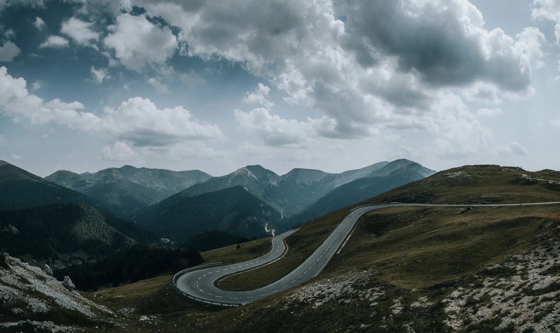 A mountain road winding down a grassy slope into a forested valley under heavy clouds, Nockalmstrasse, Austria.